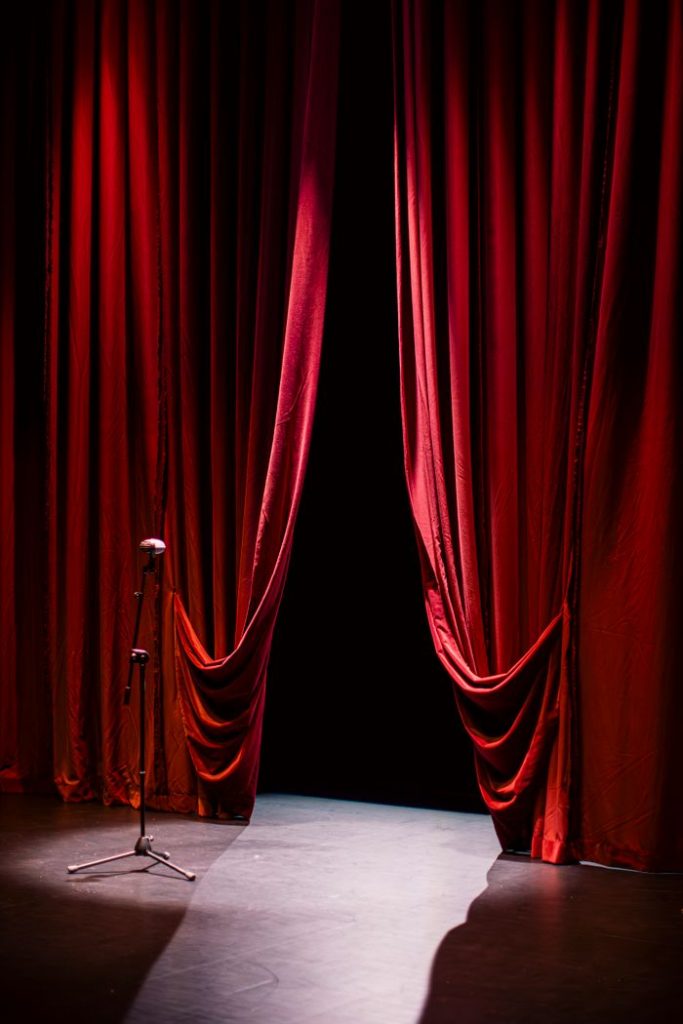 A theater stage with closed red curtains and a single microphone in the spotlight, creating a dramatic ambiance.